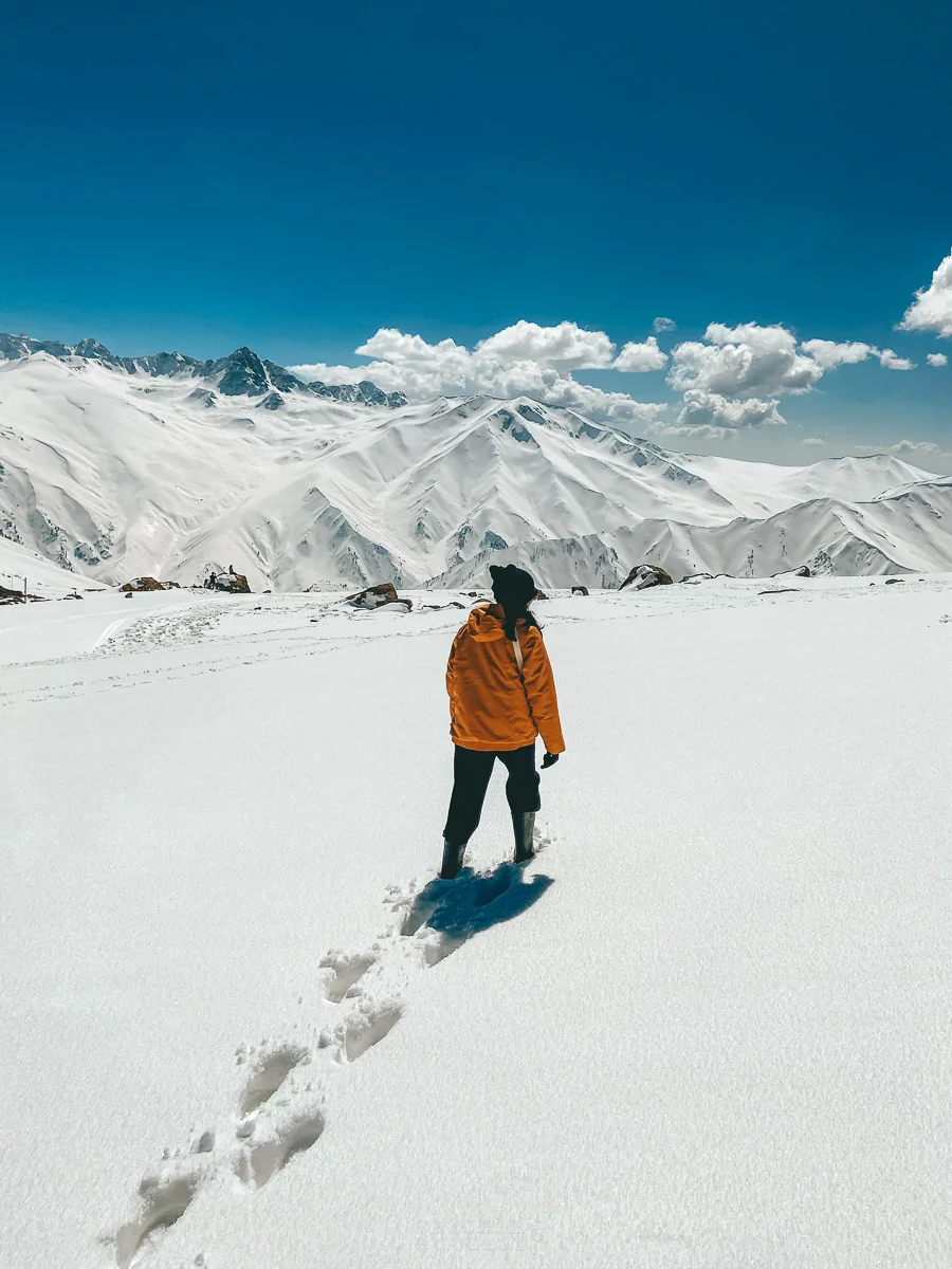 A girl wearing a yellow jacket walks on the snow facing the majestic Himalayan peaks
