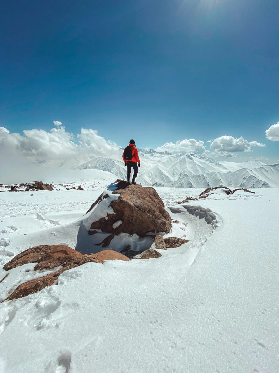 A guy wearing an orange jacket stands over a rock on top of a snowy peak looking out towards the Himalayan peaks
