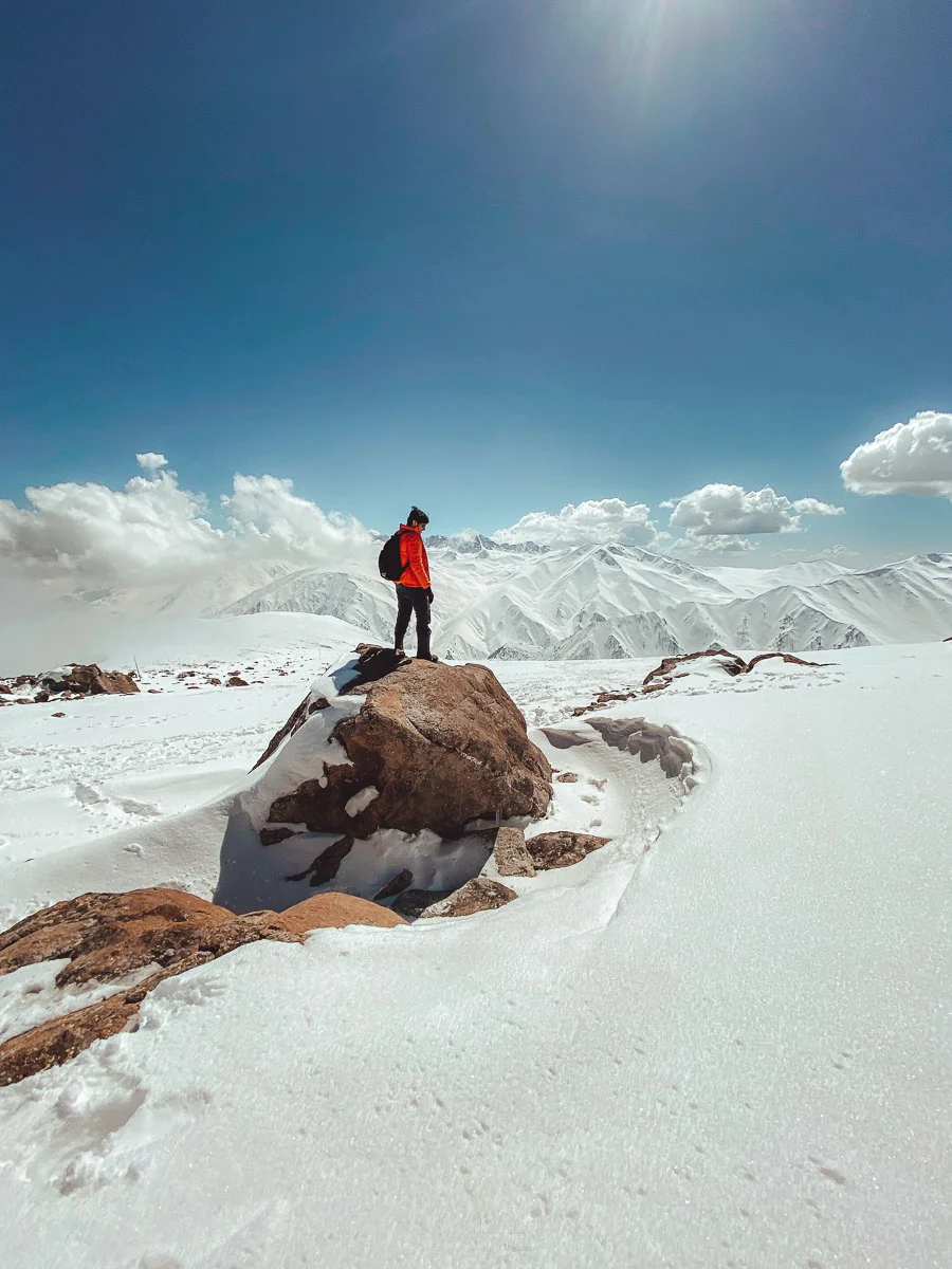 A guy wearing an orange jacket stands over a rock on top of a snowy peak looking out towards the Himalayan peaks