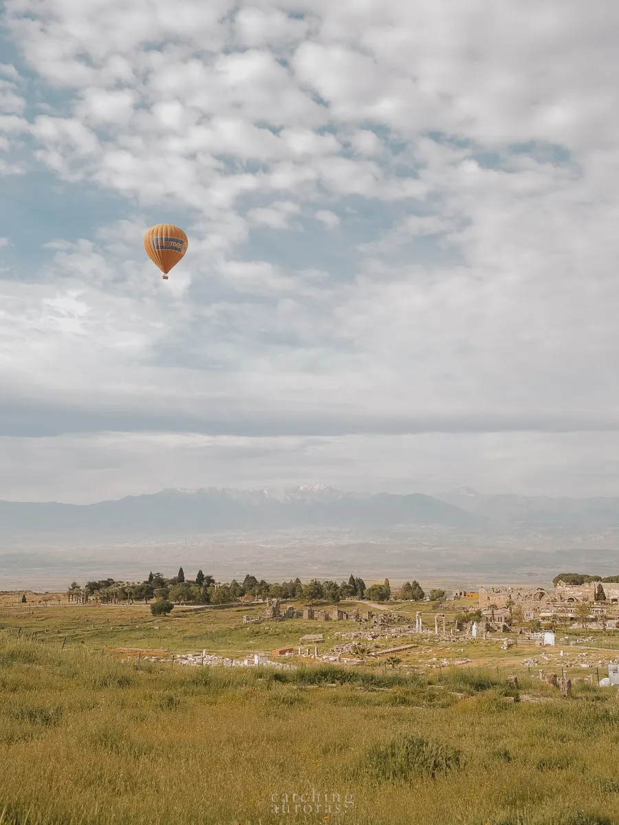 A hot air balloon flies over Hierapolis ruins interspersed between a green landscape