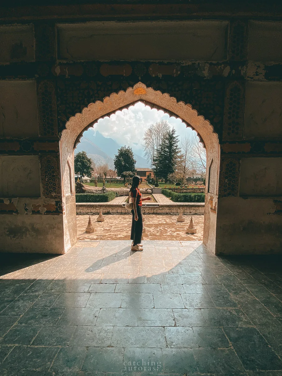 A girl wearing a red top stands silhouetted in front of a Mughal arch with a garden in the background in this Things to do in Srinagar blogpost.