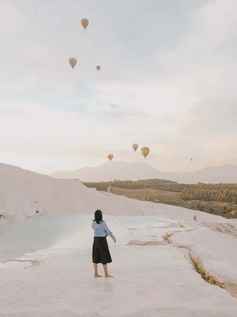 A girl stands on a white terrace with hot air balloons flying in the background