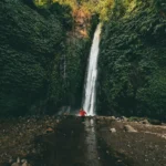 A girl wearing a red shrug stands in front of a thin and tall waterfall surrounded by lush greenery. If you are looking for a 'famous closest waterfall near me' in Munduk, this one is it.