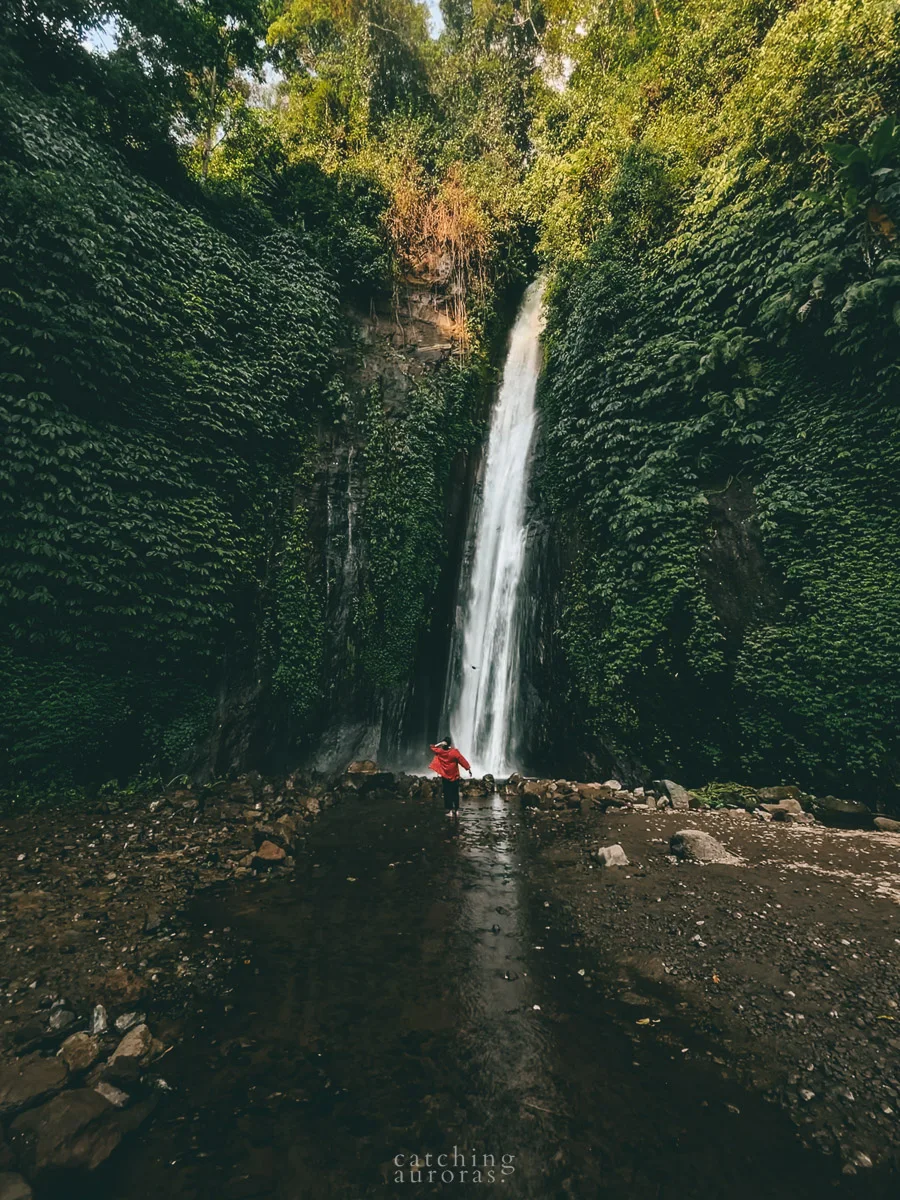 A girl wearing a red shrug stands in front of a thin and tall waterfall surrounded by lush greenery. If you are looking for a 'famous closest waterfall near me' in Munduk, this one is it.